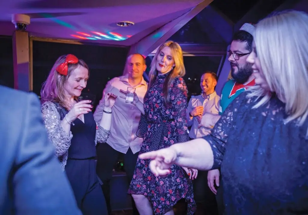 A festive Christmas Party Cruise boat sailing on the Thames at night with London landmarks illuminated, featuring a beautifully set dinner table, live entertainment, and cheerful guests